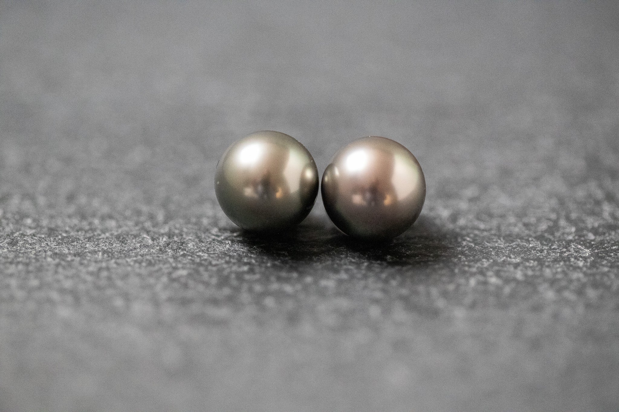 Zwei runde, graue Tahitiperlen mit intensivem Schimmer, nebeneinander auf neutralem Untergrund präsentiert
