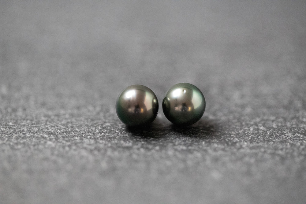 Zwei runde, dunkle Tahitiperlen mit starkem, metallischem Glanz auf dunklem Untergrund nebeneinander platziert